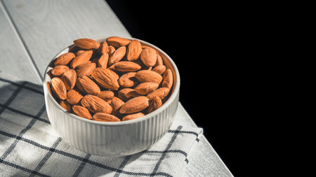 White bowl brimming with almonds resting on a checkered cloth atop a wooden table, creating a cozy,...