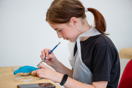 Young artist focused on painting a decorative object with precision. Wearing a protective apron, she uses a brush to add vibrant colors, showcasing creativity and concentrationの写真素材