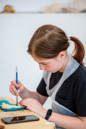 Young artist focused on painting a decorative object with precision. Wearing a protective apron, she uses a brush to add vibrant colors, showcasing creativity and concentrationの写真素材