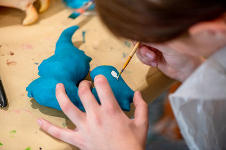 Female designer painting a small blue dinosaur model with white paint using a small brush, working on a cardboard covered table in a design studioの写真素材