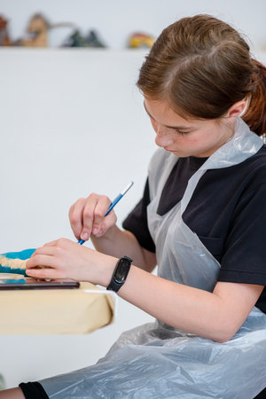 Young artist focused on painting a decorative object with precision. Wearing a protective apron, she uses a brush to add vibrant colors, showcasing creativity and concentrationの写真素材