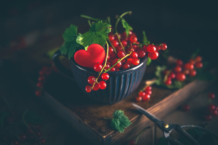 Freshly picked red currant berries and leaves are overflowing a green cup decorated with a red heart, sitting on a wooden box with gardening shears nearbyの写真素材