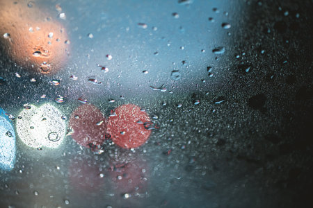 Raindrops on a window pane create a textured surface, distorting colorful street lights and city activity into soft, out of focus background bokeh, capturing a moody urban atmosphere