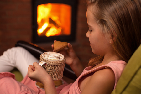 Young girl sitting in front of fireplace with a hot chocolate and cookies - relaxation concept, closeup, shallow depthの写真素材