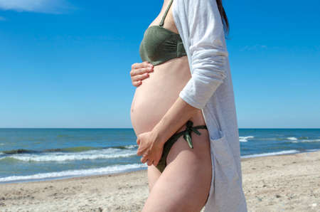A pregnant young woman in anticipation of a baby stands hugging her belly on the seashoreの写真素材