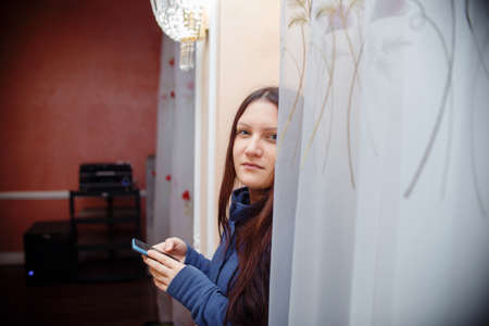 girl in a blue sweater in a bright room standing by the window, holding a phone and looking at the...