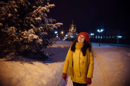 the girl's portrait on a city footpath, in the winter during snowfall, against the background of church, a magnificent fir-tree and a number of lamps. the evening lit with blue firesの写真素材
