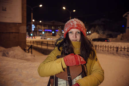 the girl's portrait against the background of the evening city which to something is surprised and holds in the winter a backpack in hand. in the light of city lamps.の写真素材
