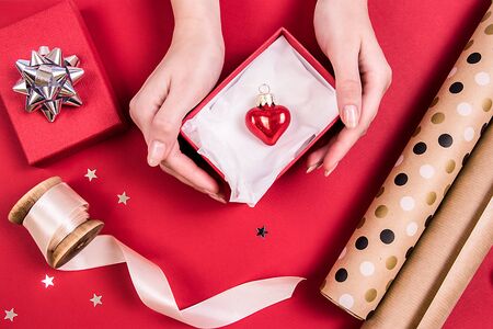 Red glass heart in a red box, beige ribbon, design paper. Top view. Holidays concept.