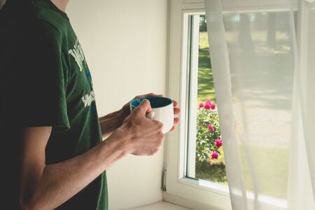 Man with a cup in t-shirt near a window. Morning time. Relax.の写真素材