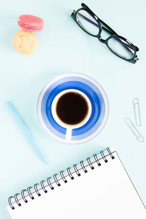 Macaroons, a cup of black coffee, an open note with white page, pen, glasses on a pastel blue background. Image for your design.の写真素材