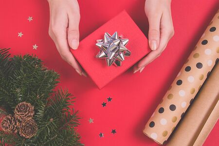 Woman's hands holding a red gift with a silver bow on a red background. Christmas tree wreath,...