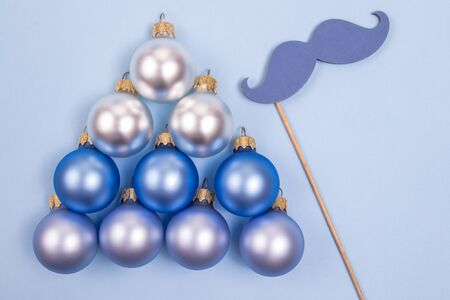 Blue fake mustache and matte blue baubles laid in a shape of Christmas tree on a pastel blue background. Christmas and New Year concept.の写真素材