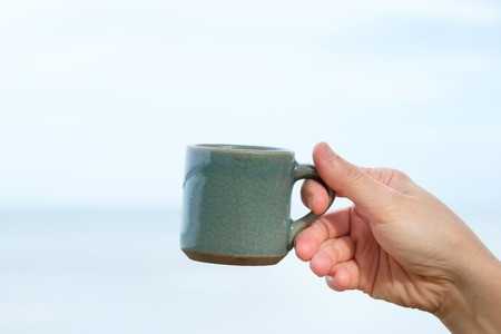 Hand holding ceramic cup of coffee on sea and sky background, with peace and joy conceptの写真素材