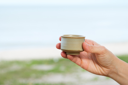 Hand holding ceramic cup of coffee on sea and sky background, with peace and joy conceptの写真素材