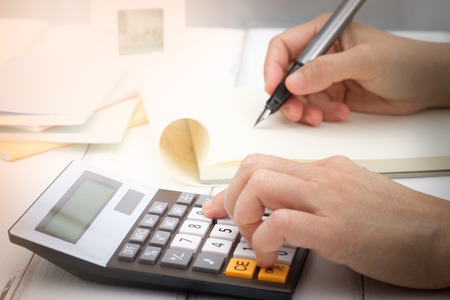Hand writing and counting on calculator in office on wood desk about financial data analyzing, Choose focus point.の写真素材