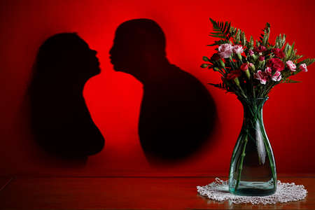 Concept love, valentine's day, passion. Copy space. Image of a green vase with roses and blossoms. Red background with shadows of a man and a woman kissing.の写真素材