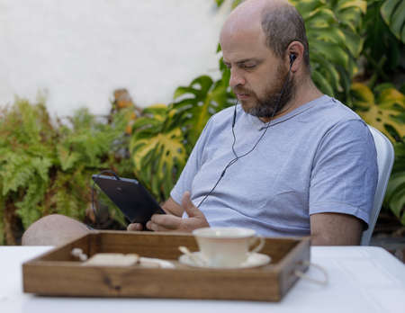 A man is staring intently at a digital device, listening headphones in his ears. The person may be...