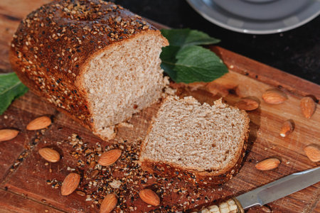 Fresh homemade whole wheat bread, with mixed cereals of seeds and grains; on wooden board. Accompanied by a rustic knife, almonds and mint leaves.の写真素材