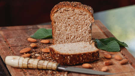 Fresh homemade whole wheat bread, with mixed cereals of seeds and grains; on wooden board. Accompanied by a rustic knife, almonds and mint leaves.の写真素材