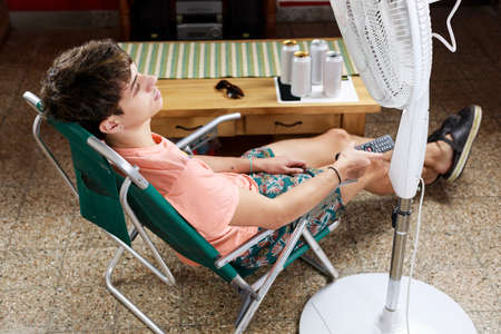 Man in the living room of his house, lying on a beach chair, near the fan. Concept of summer at home, vacation, relaxation, rest.の写真素材