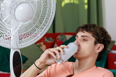 A Concept of summer at home. Young man on a beach chair, drinking beer or soft drink, near the fan.の写真素材