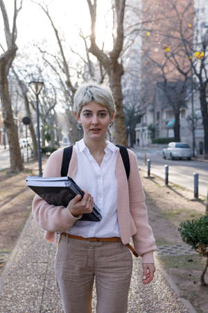 A Front view of young Latina student walking down city street on a sunny autumn day. She is carrying books and folders.の写真素材