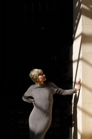 Young woman model, blonde and short hair. Wearing a dress. Standing in front of an old building. Leaning against the wall. Looking up. Urban exterior in daytime.の写真素材