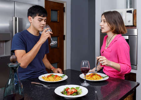 Young couple in the kitchen of their home, have dinner and drink red wine while chatting.