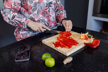 Caucasian womans hands chopping bell peppers on a wooden board on the kitchen counter. On the side the smartphone.の写真素材