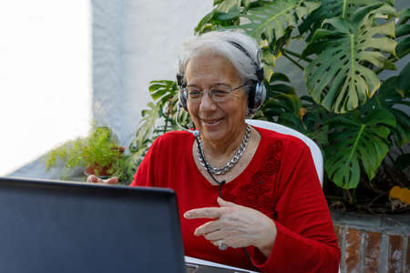 Happy grandmother looks at laptop camera, wears headphones, talks to family or communicates with friends, via video call. Older adult communicating with modern technology.の写真素材