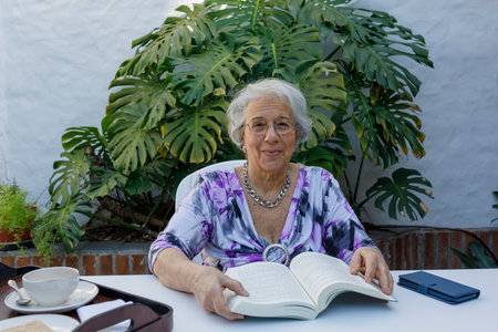 Grandmother in the courtyard of her house reading a book while having breakfast. She looks at the camera smiling. Quality leisure time. Lifestyle of older adults. Retirement.の写真素材