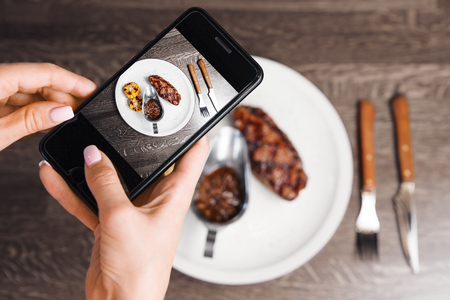 Photo on smartphone of beefsteak with grilled corn and sauce in iron bowl on white plate, closeup view on just prepared dish with meat and vegetablesの写真素材