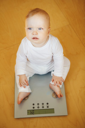Nice baby with big grey eyes and slightly surprised facial expression sits on modern electronic scales on floorの写真素材