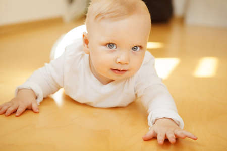 Adorable baby with big light eyes and blond hair in white crawlers lies on wooden floor with calm face expression and looks directly in camera.の写真素材