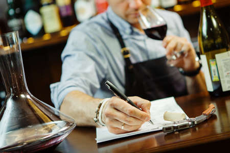 Male sommelier tasting red wine and making notes at bar counter. Bottle of wine nearby. Professional expert appreciates quality of alcoholic beverage, degustation processの写真素材