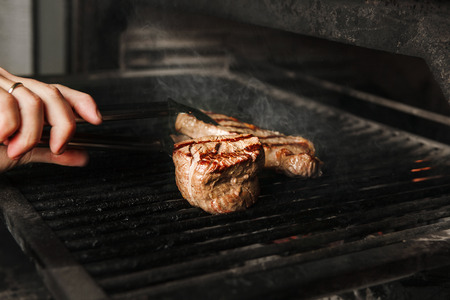 Beef steak medium roast cooking on grill, person turns the meat with tongs on lattice, preparing main course in restaurant closeup viewの写真素材