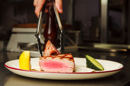 Man holds by tongs medium rare beef steak on white plate with lemon and cucumber closeup view on well done meal just from stoveの写真素材