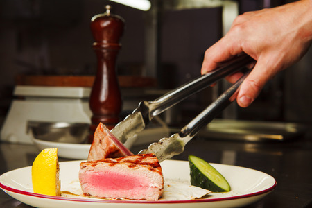 Man holds by tongs medium rare beef steak on white plate with lemon and cucumber closeup view on well done meal just from stoveの写真素材