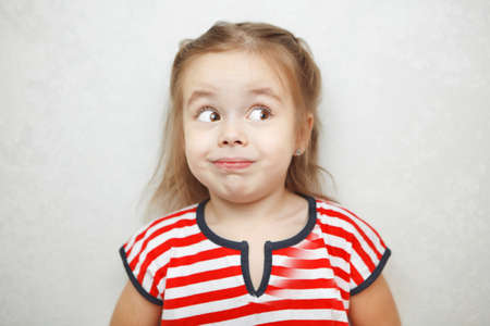 Surprised little girl with arched eyebrows and weird smile looks left portrait photo. Funny child in striped T-shirt with bright facial expression that shows astonishment.の写真素材