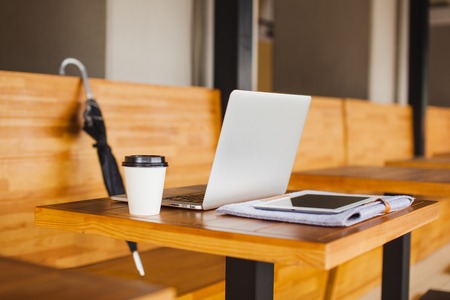 Modern equipment on wooden table at cafe. Open laptop, tablet on folder with documents, glass of coffee and closed umbrella on background.の写真素材