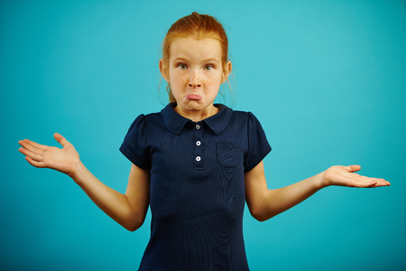 Comical girl with freckles and red hair expresses puzzlement or ignorance, lifted her shoulders, spreads hands to the side, does not possess information, demonstrates confusion, on blue isolated.の写真素材
