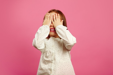 Little girl covered her eyes with hands and smiles on pink isolated background.の写真素材