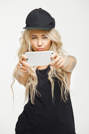 Vertical portrait of cute woman with blond hair dressed in look of swag makes selfie on phone over white isolated.の写真素材