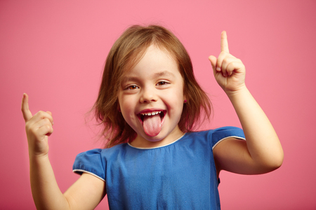 Cheerful little girl dancing with her hands raised and tongue sticking out, portrait of joyful female child over pink background.の写真素材