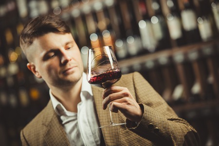 Sommelier consider red wine in bokal on background of shelves with bottles in cellar. Male appreciating color, smell, quality and sediments of drink.の写真素材