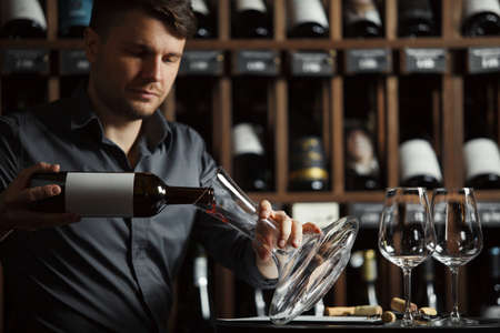 Serious sommelier pouring red wine from opened bottle in decanter designed to degustate and taste alcohol beverages, cellar full with containers collection having labels placed on shelvesの写真素材