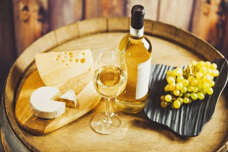 White wine in glass, bottle with grape and cheese on barrel front wood wall background. Degustation, drink, alcohol beverage.の写真素材