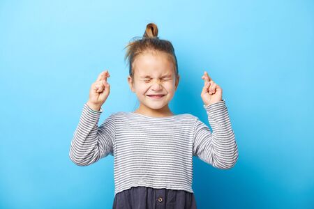 Closeup beautiful child girl crossing her fingers, she squeezed her eyes shut, has a hope, isolated blue background.の写真素材