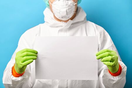 Close up of medical worker in protective sterile clothing and respiratory mask stands on blue background with white sheet for text. Doctor makes recommendations about the spread of the virus.の写真素材